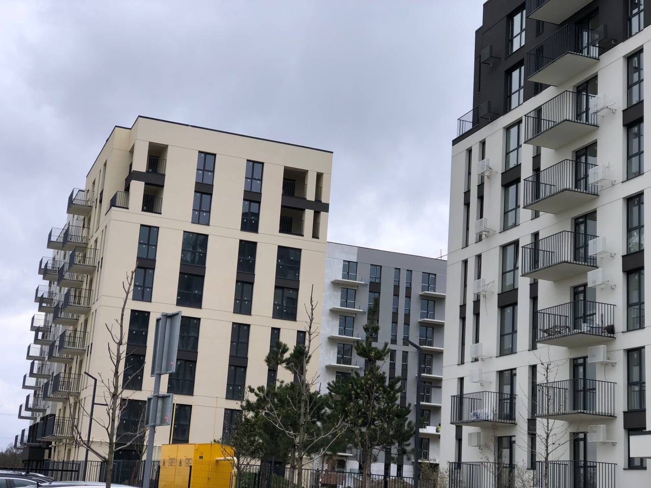 Row of modern multi-story apartment buildings with balconies on a cloudy day; yellow utility box and young trees in foreground.