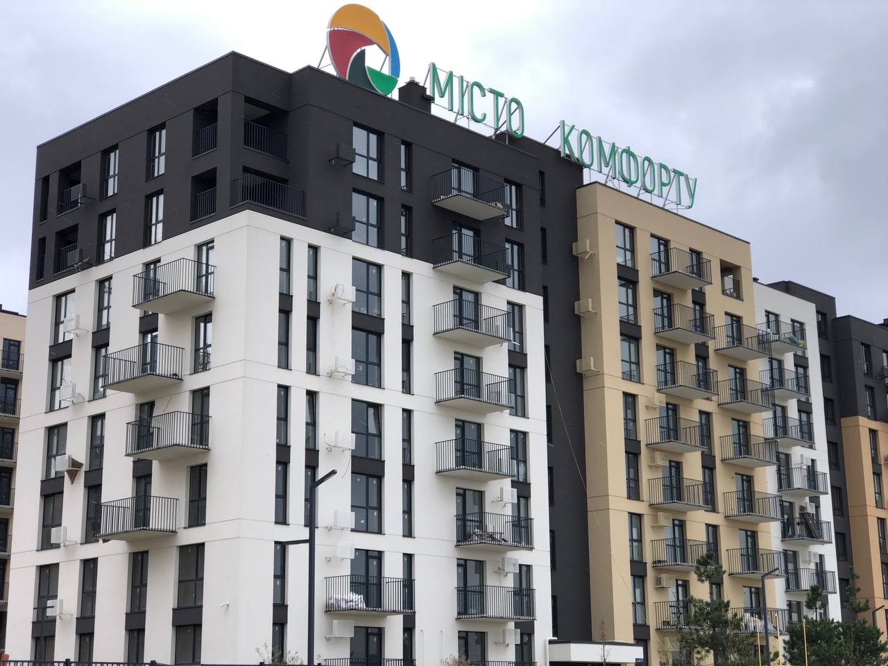 Modern multi-story apartment building with white, black, and beige façades and balconies along each wing.