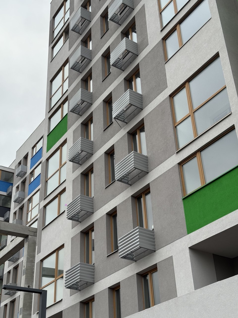 Modern gray apartment building with numerous small metal balconies and large windows, green and blue accent panels on the facade.