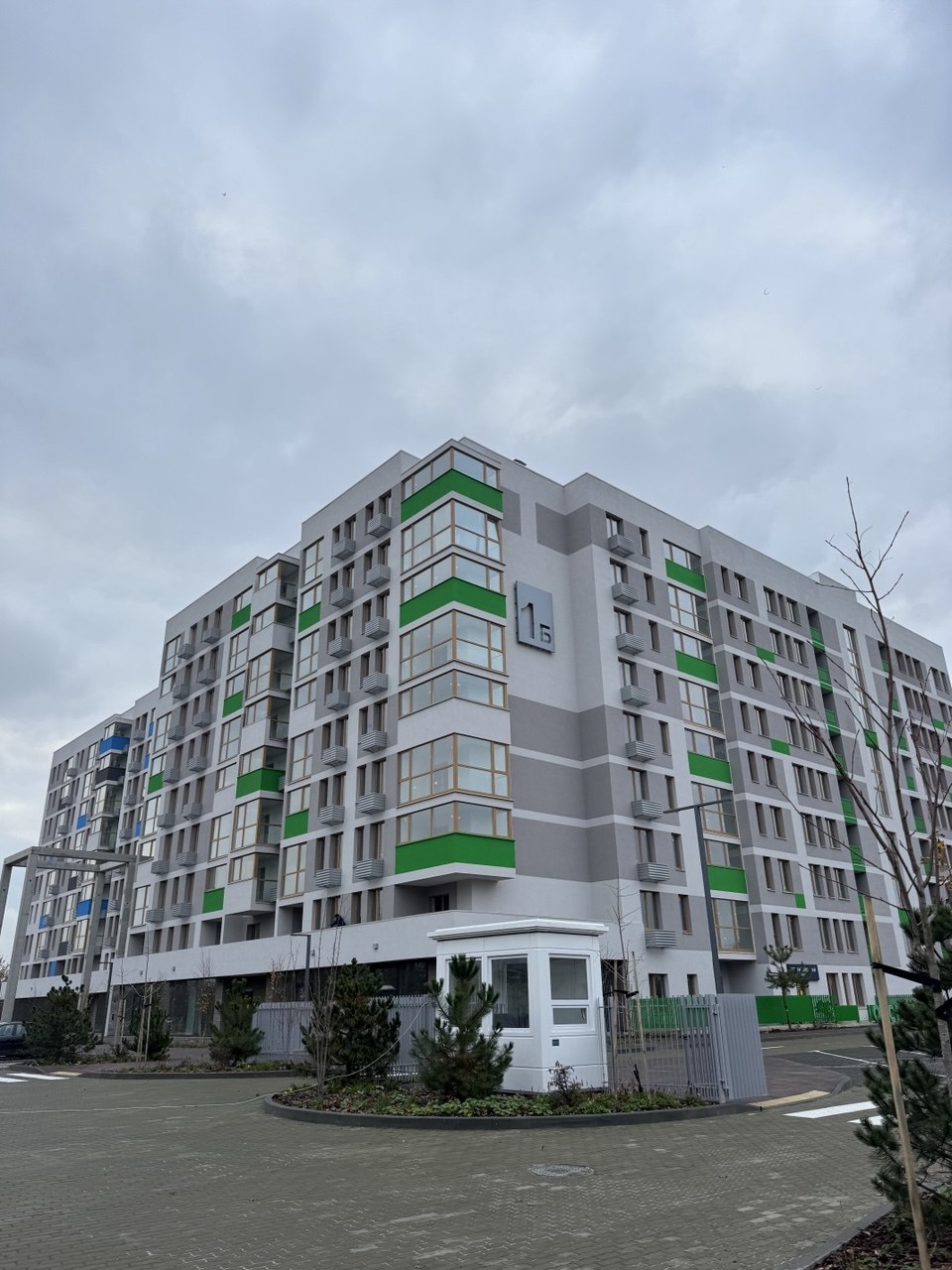 Six-story apartment building with gray facade and bold green vertical accents, viewed from the circular driveway area at the entrance.