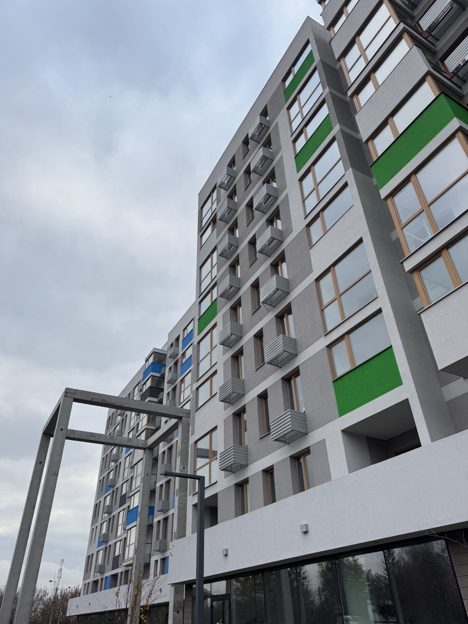 Modern multi-story apartment building with gray facade and green/blue panel accents, balconies, and large ground-floor windows.