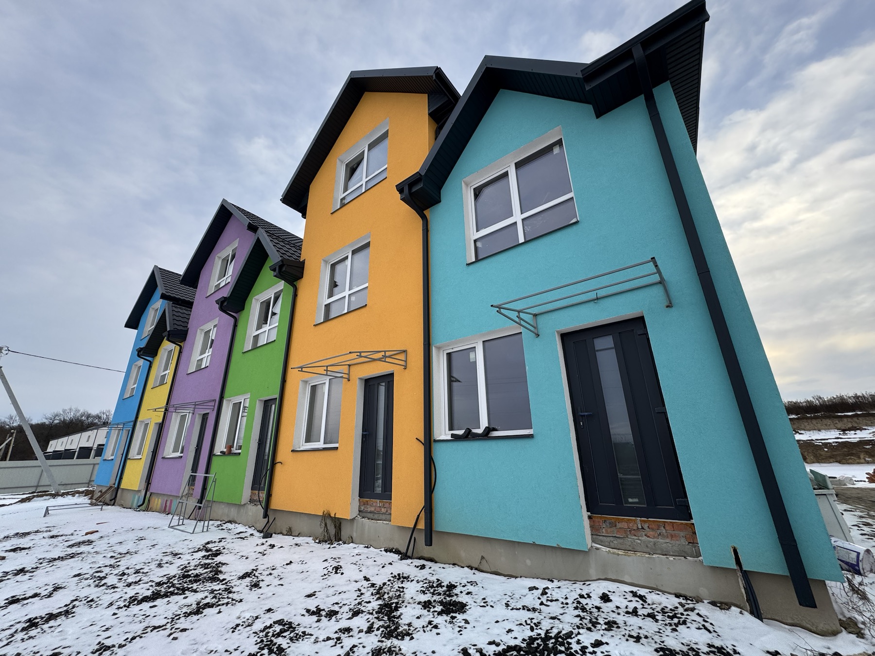 Row of colorful houses in blue, purple, yellow, green, orange, and turquoise, with white-framed windows and dark doors, under a cloudy sky and snowy ground.