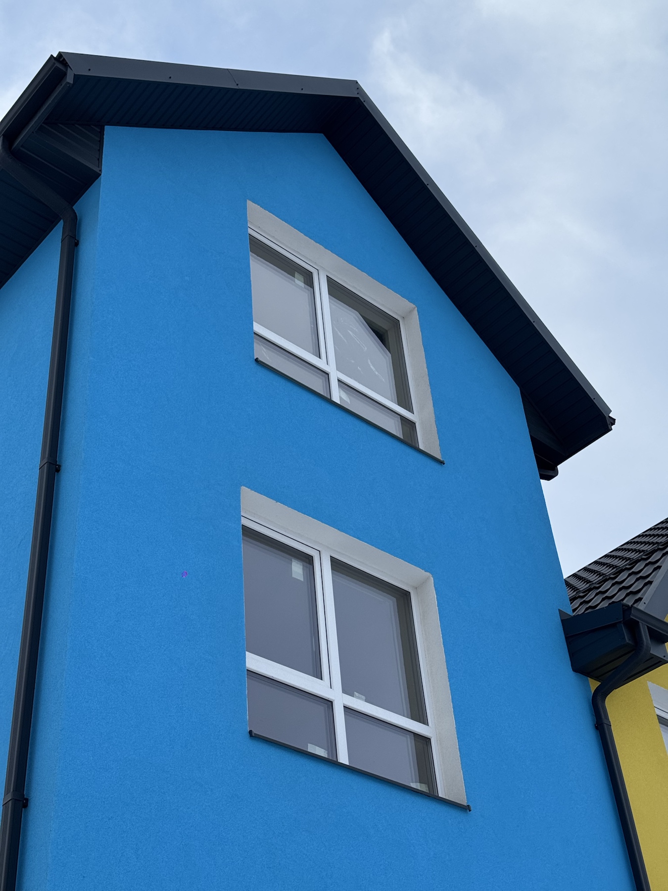 Blue house facade with two white-framed windows, gabled dark roof, and black gutter along the left side under a cloudy sky.
