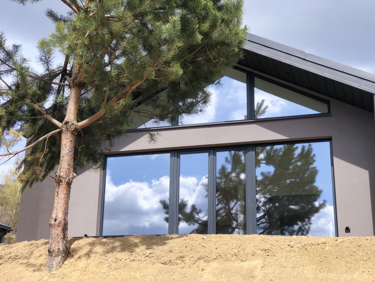 Modern house with large glass doors reflecting a blue sky and trees, built on a dirt slope with a pine tree leaning over the facade