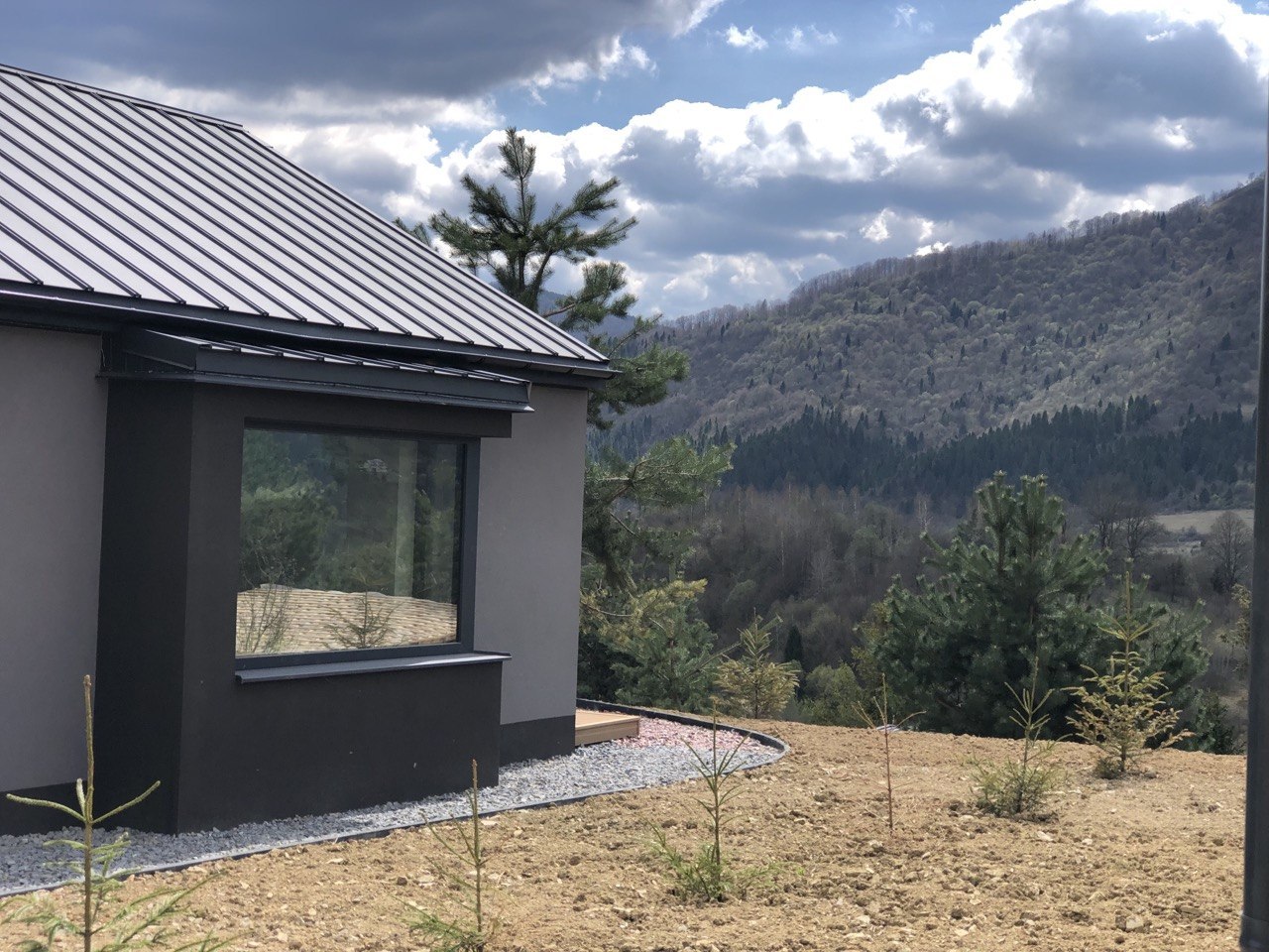 Modern grey house with a metal roof on a hillside, a large window facing a forested mountain landscape.