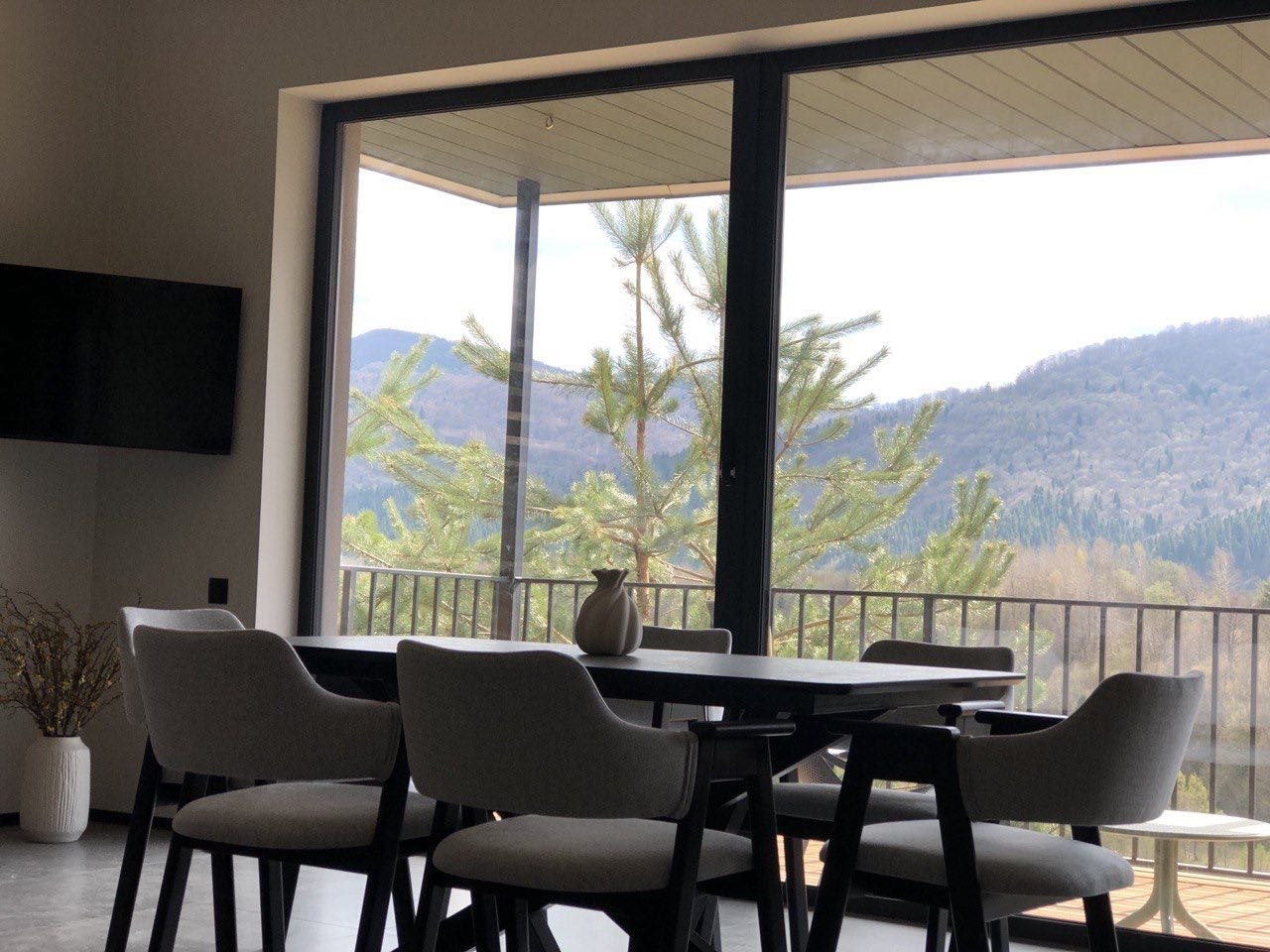 Dining area with a dark table and gray chairs by a large glass wall showcasing a mountain landscape in the distance.