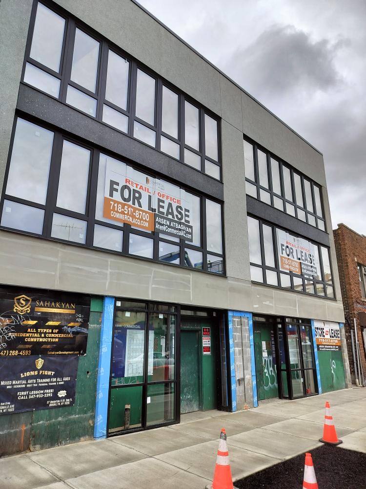 Three-story commercial building with large windows and two signs reading 'FOR LEASE' on the upper floor; storefronts below with glass doors and construction cones on the sidewalk.