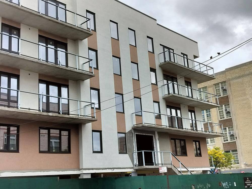 Modern multi-story residential building with balconies under construction and a green fence in foreground.